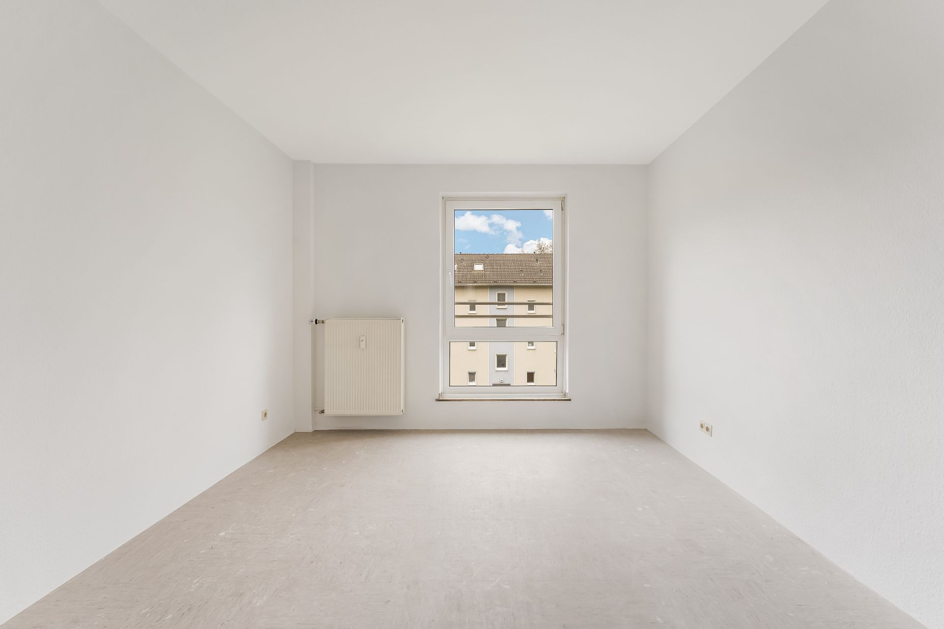 Leerer, heller Raum mit Fenster, Heizung und Blick auf ein gegenüberliegendes Gebäude.