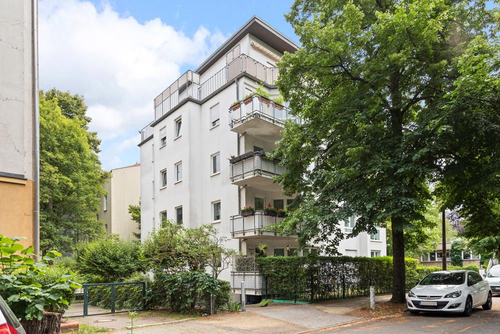 Mehrstöckiges Wohnhaus mit Balkonen, Bäumen und geparktem Auto an der Straße.