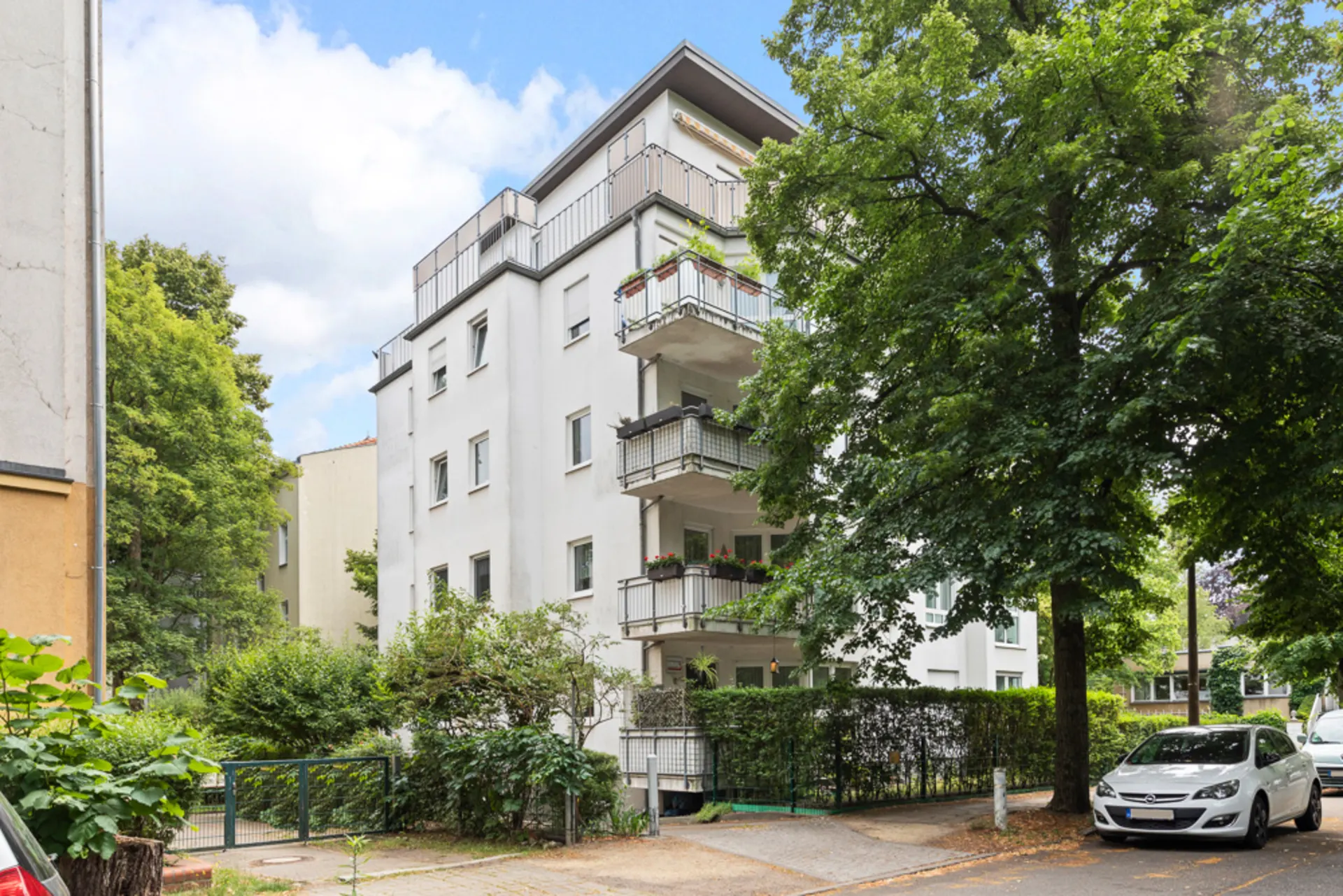 Mehrstöckiges Wohnhaus mit Balkonen, Bäumen und geparktem Auto an der Straße.