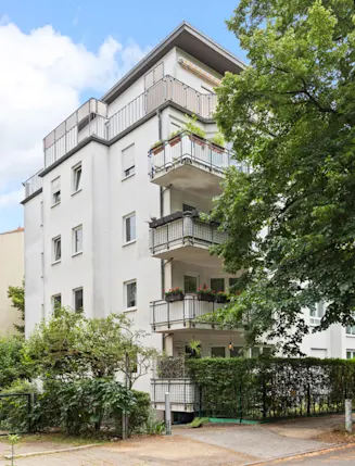 Mehrstöckiges Wohnhaus mit Balkonen, Bäumen und geparktem Auto an der Straße.