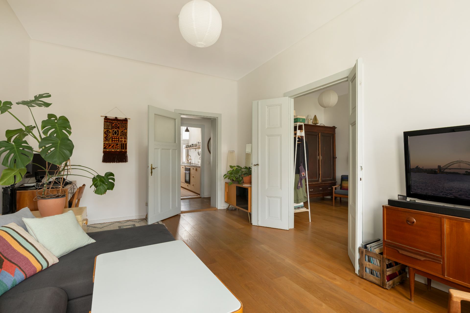 Helles Wohnzimmer mit Holzboden, Sofa, Fernseher, Pflanzen und geöffneten Türen zu anderen Räumen.