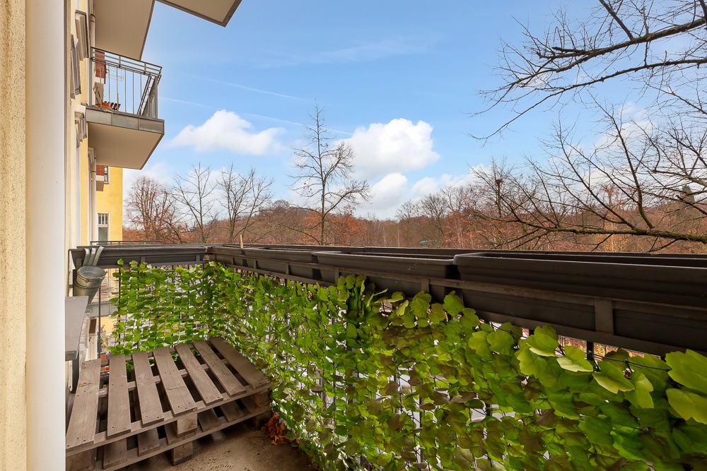 Balkon mit Holzpalette und grünem Sichtschutz, im Hintergrund Bäume und blauer Himmel.