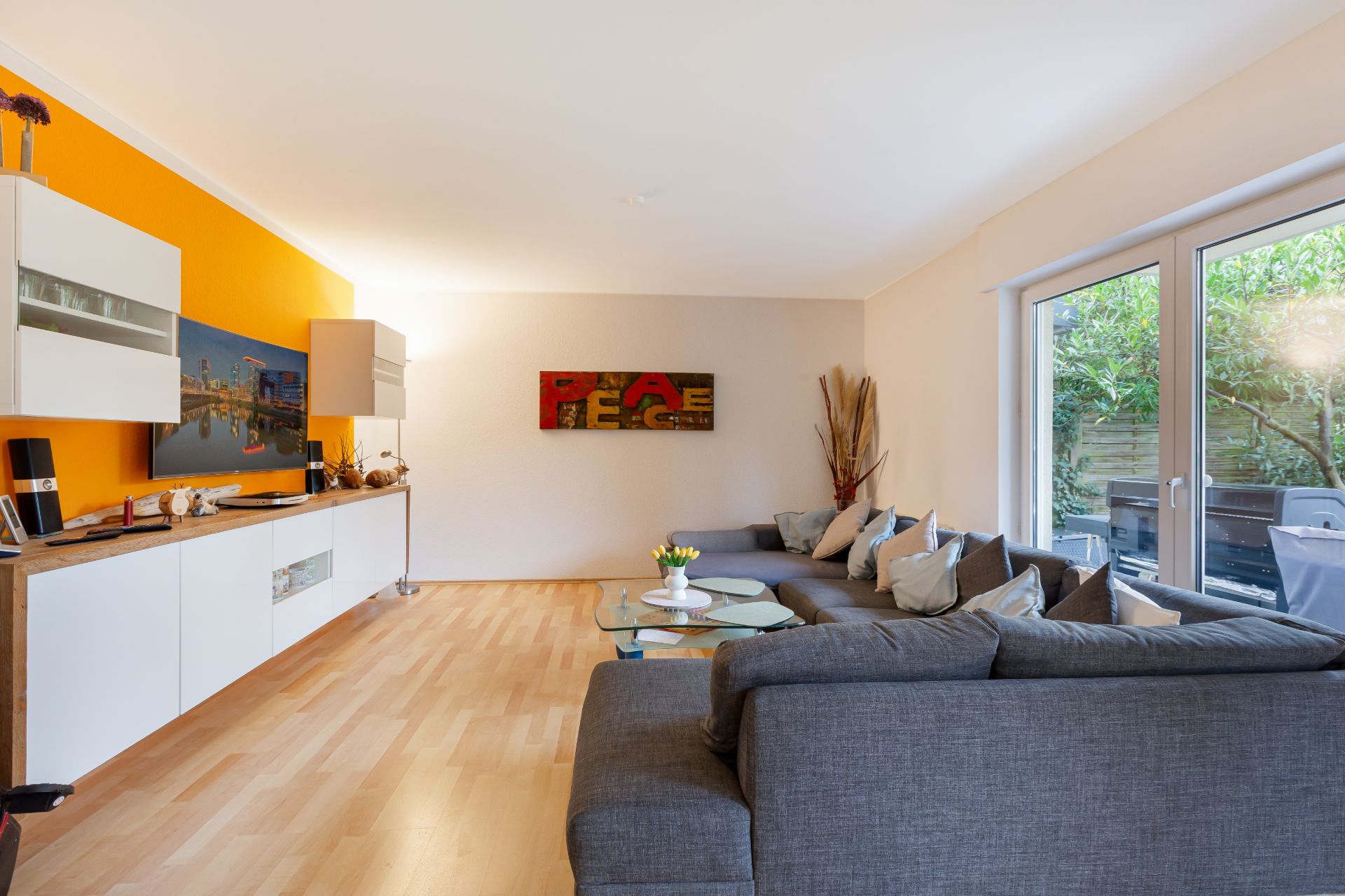 Helles Wohnzimmer mit grauem Sofa, Glastisch, gelber Wand und großem Fenster zum Garten.