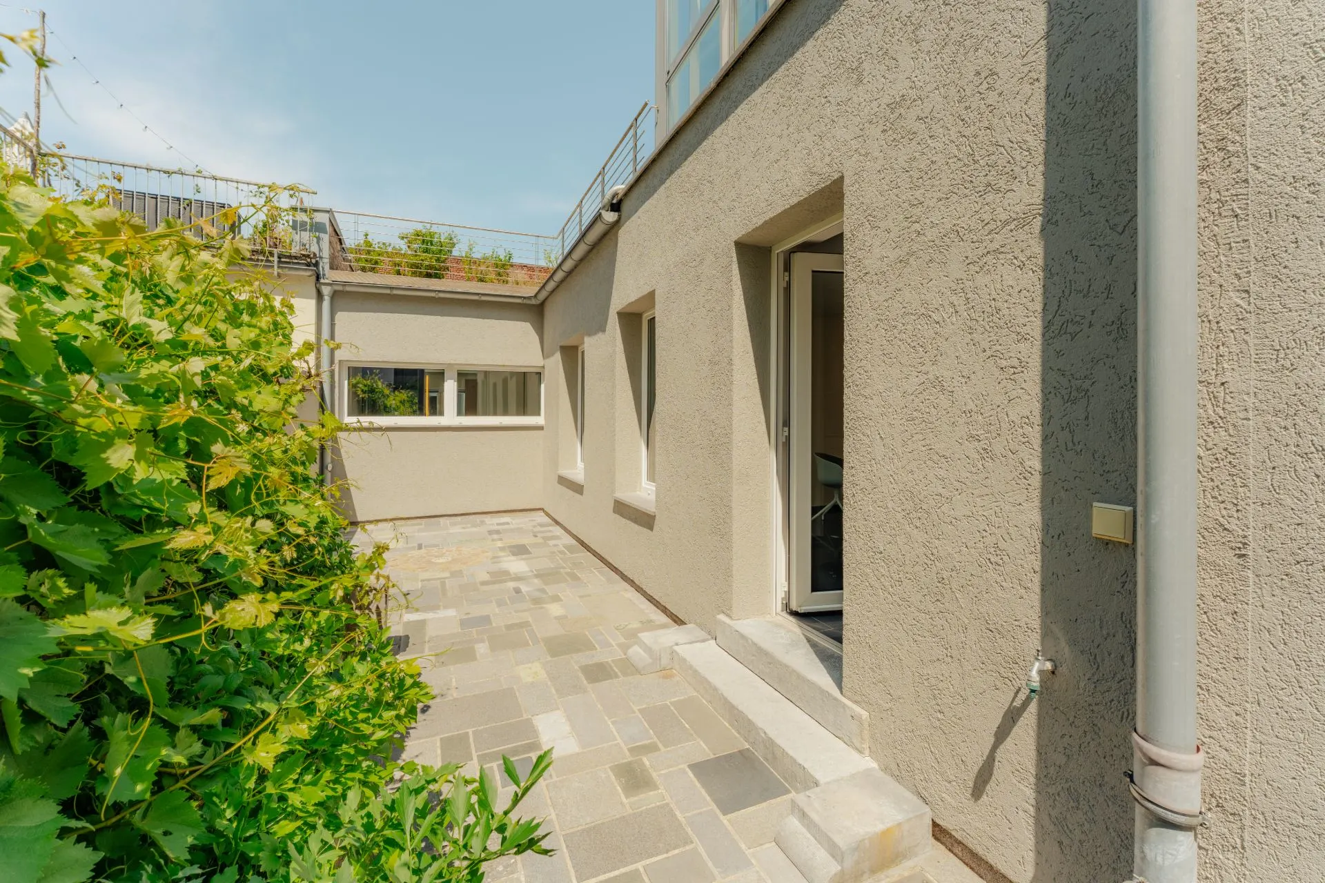 Gepflasterte Terrasse neben einem hellen Wohnhaus mit offenstehender Tür und Büschen.