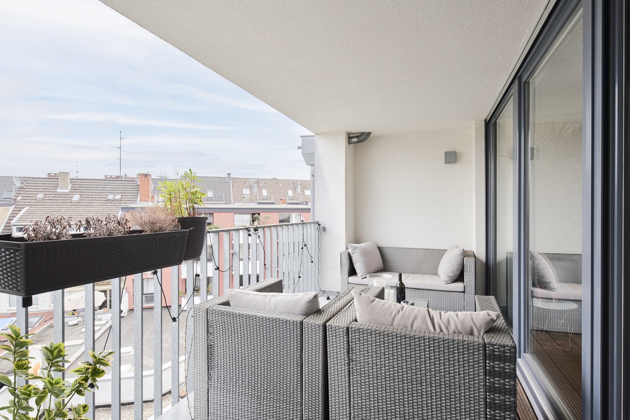 Überdachter Balkon mit grauen Polstermöbeln, Pflanzenkästen und Blick auf Nachbarhäuser.