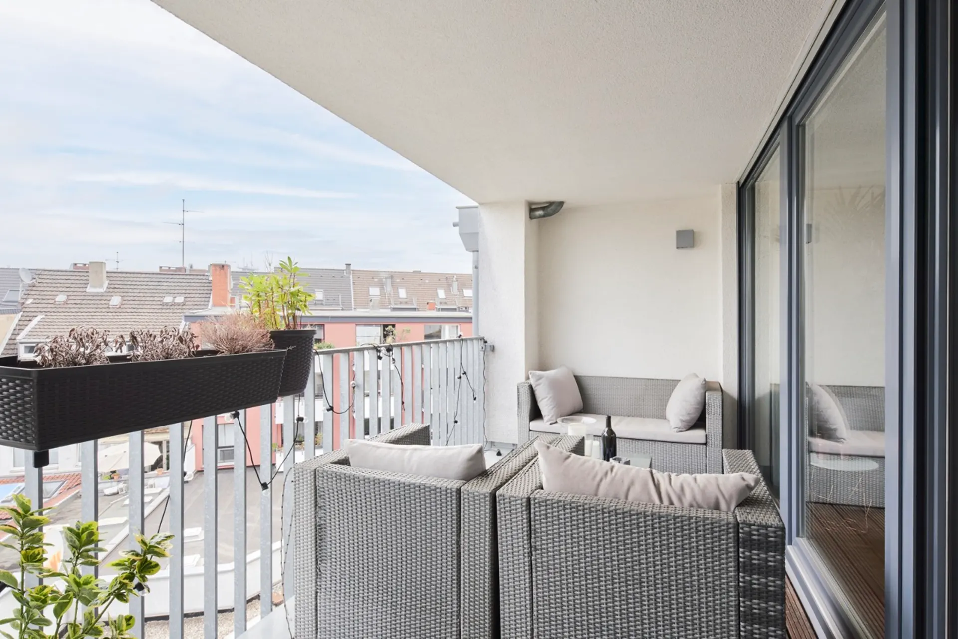 Überdachter Balkon mit grauen Polstermöbeln, Pflanzenkästen und Blick auf Nachbarhäuser.