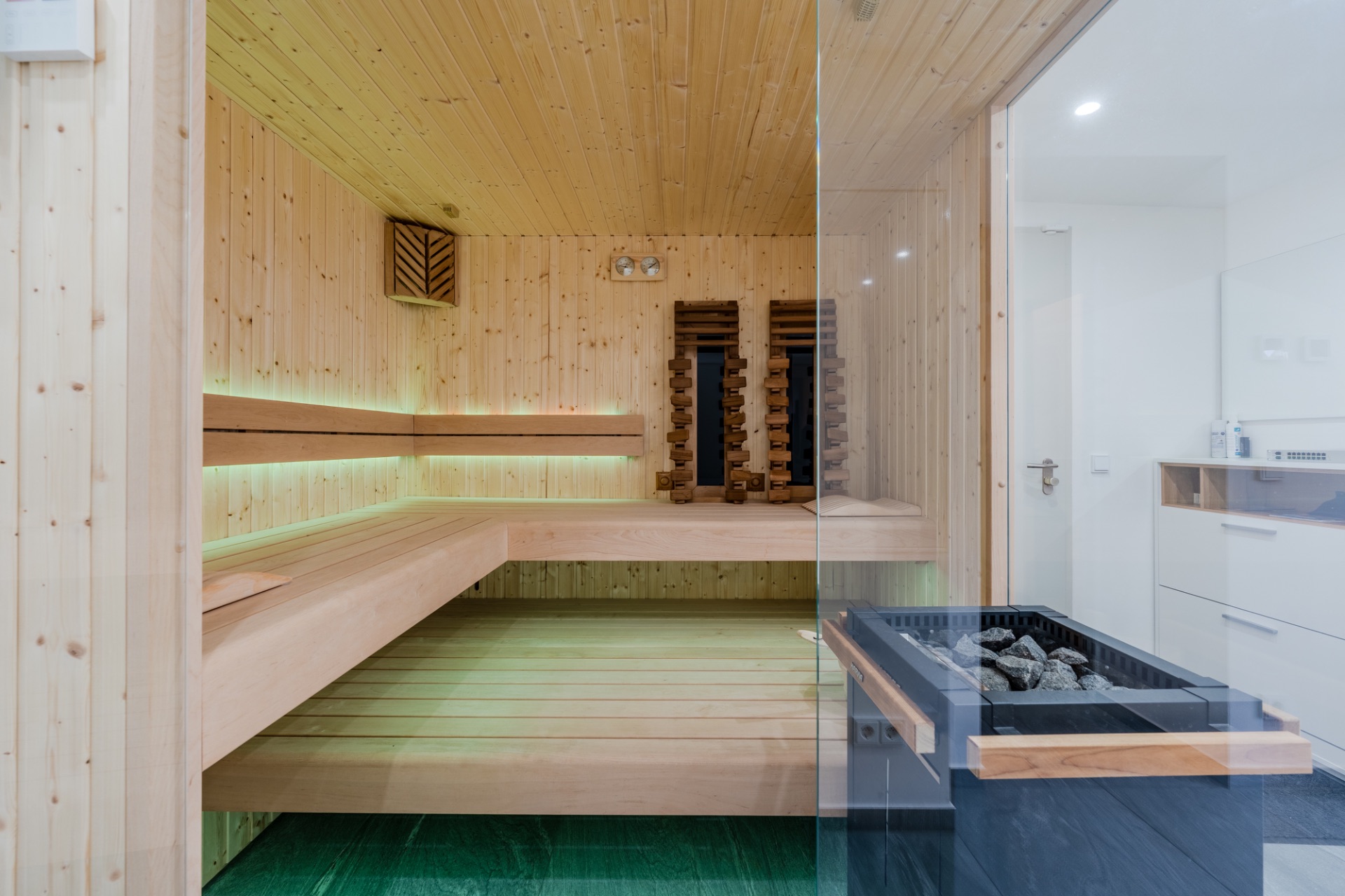 Moderne Sauna mit Holzbänken und Glastür, im Hintergrund heller Raum mit Waschbecken.