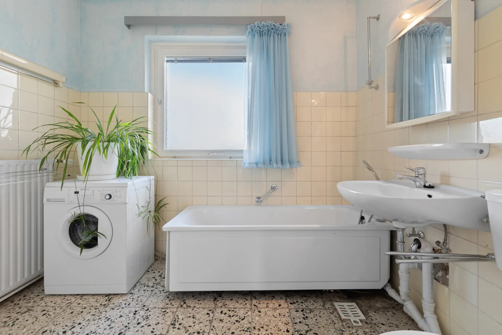 Helles Badezimmer mit Badewanne, Waschmaschine, Waschbecken und Pflanze am Fenster mit blauen Vorhängen.