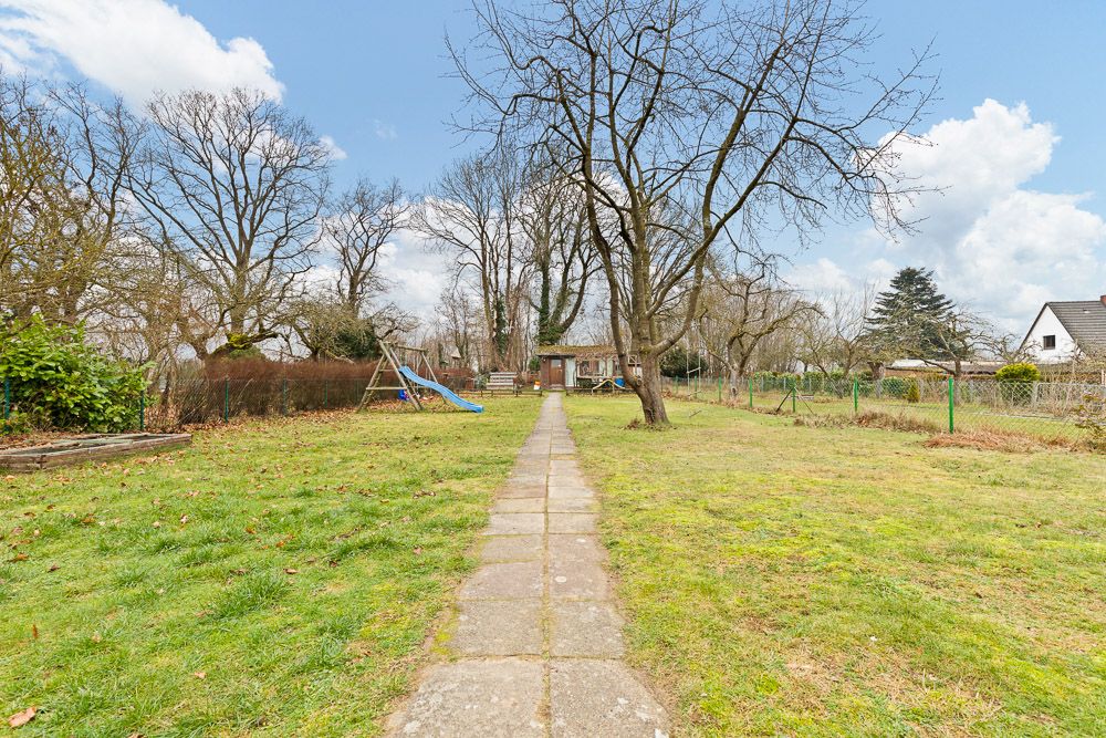 Garten mit Steinweg, Bäumen und einer blauen Rutsche, im Hintergrund ein kleines Haus.