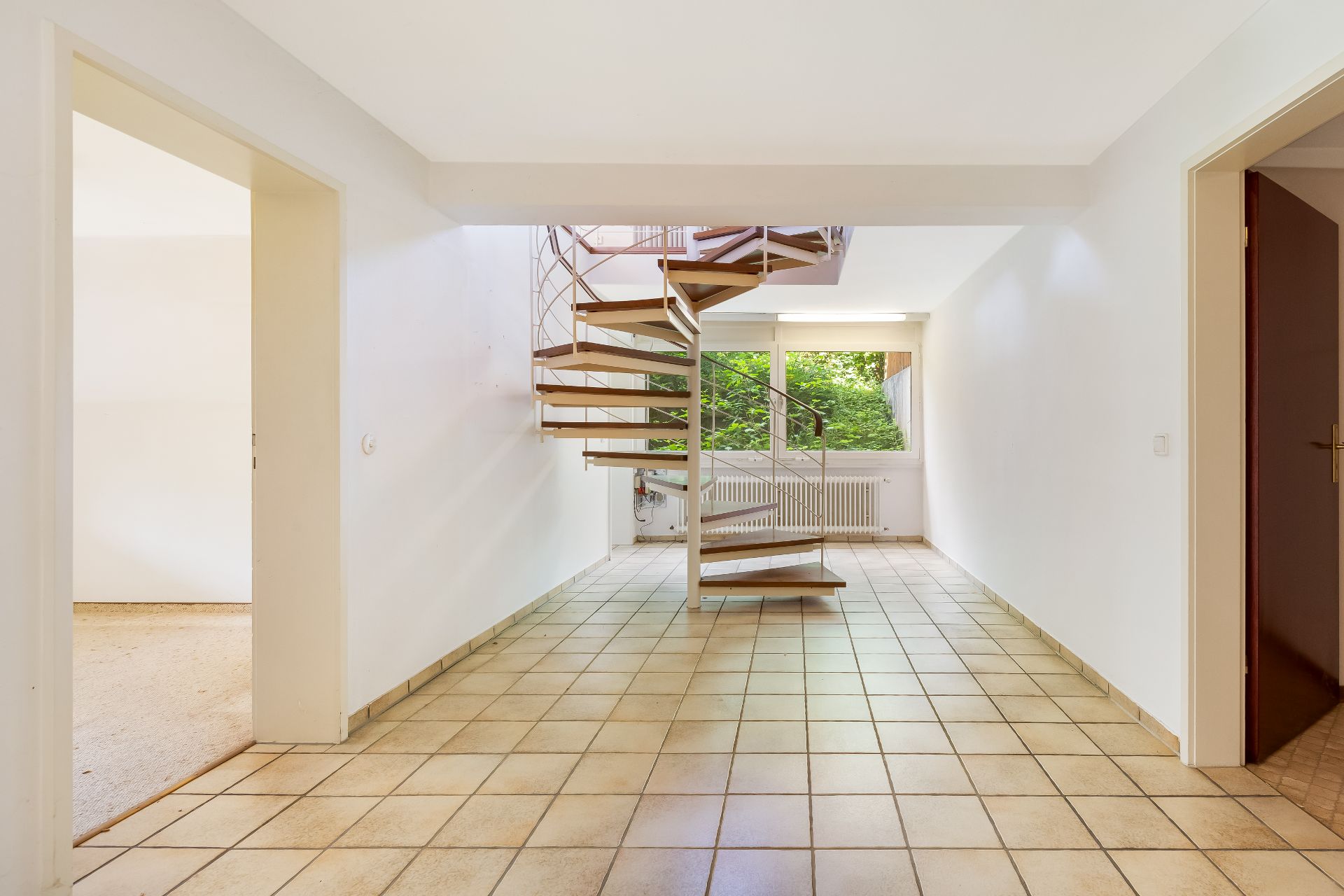 Helle Diele mit Fliesenboden und einer offenen Wendeltreppe, Fenster mit Blick ins Grüne.