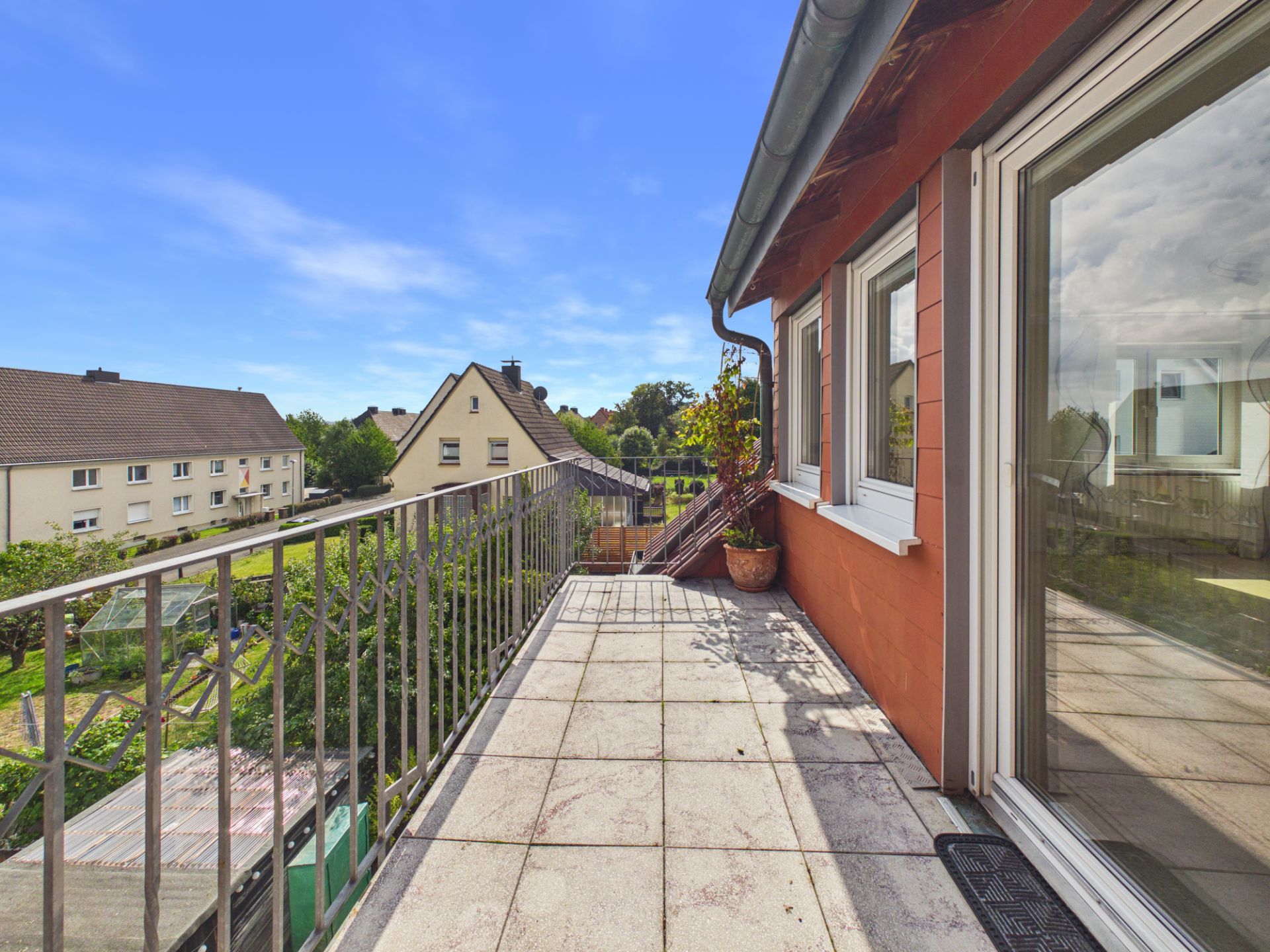 Balkon mit Fliesenboden, Geländer und Blumentopf, Blick auf Wohnhäuser und Gärten.