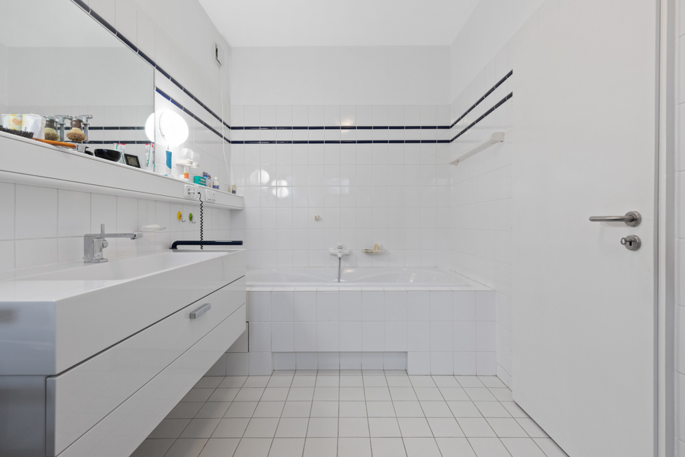 Modernes Badezimmer mit weißer Fliesenwand, Badewanne und breitem Waschbecken mit Spiegel.