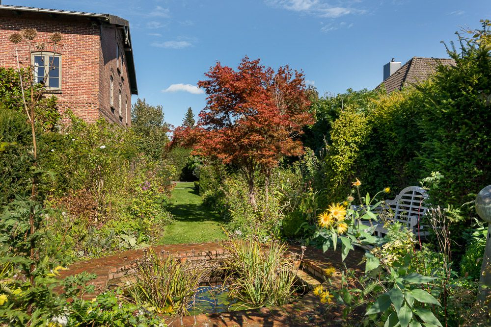 Grüner Garten mit Teich, Bank und rotem Baum neben einem Backsteinhaus.