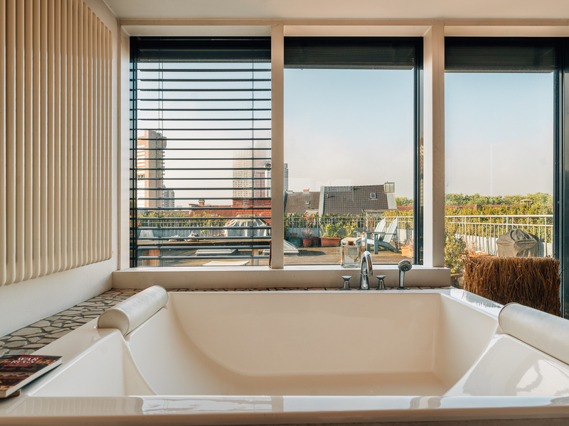 Große Badewanne vor einem Fenster mit Blick auf eine Terrasse und Stadtgebäude.