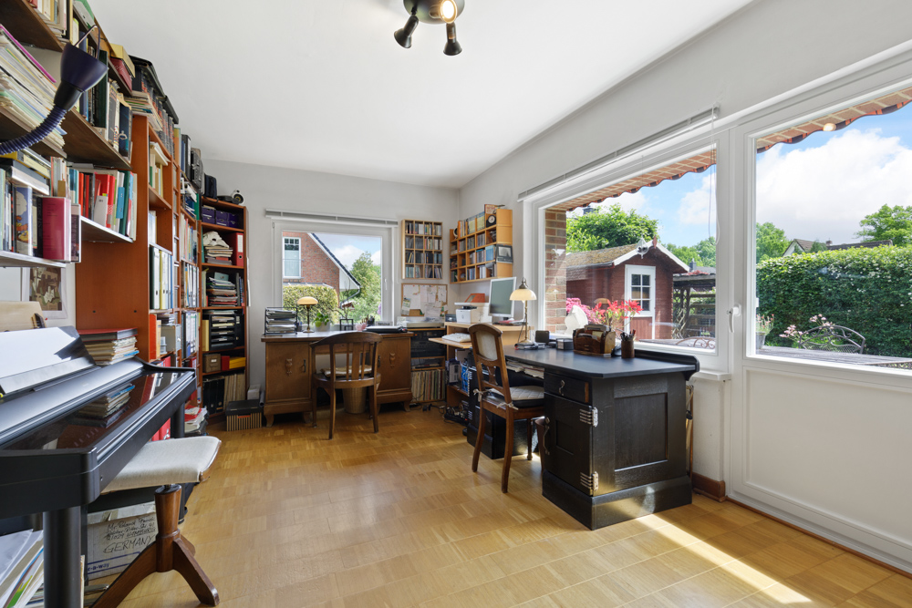 Helles Arbeitszimmer mit Schreibtischen, Regalen voller Bücher und großen Fenstern mit Blick in den Garten.