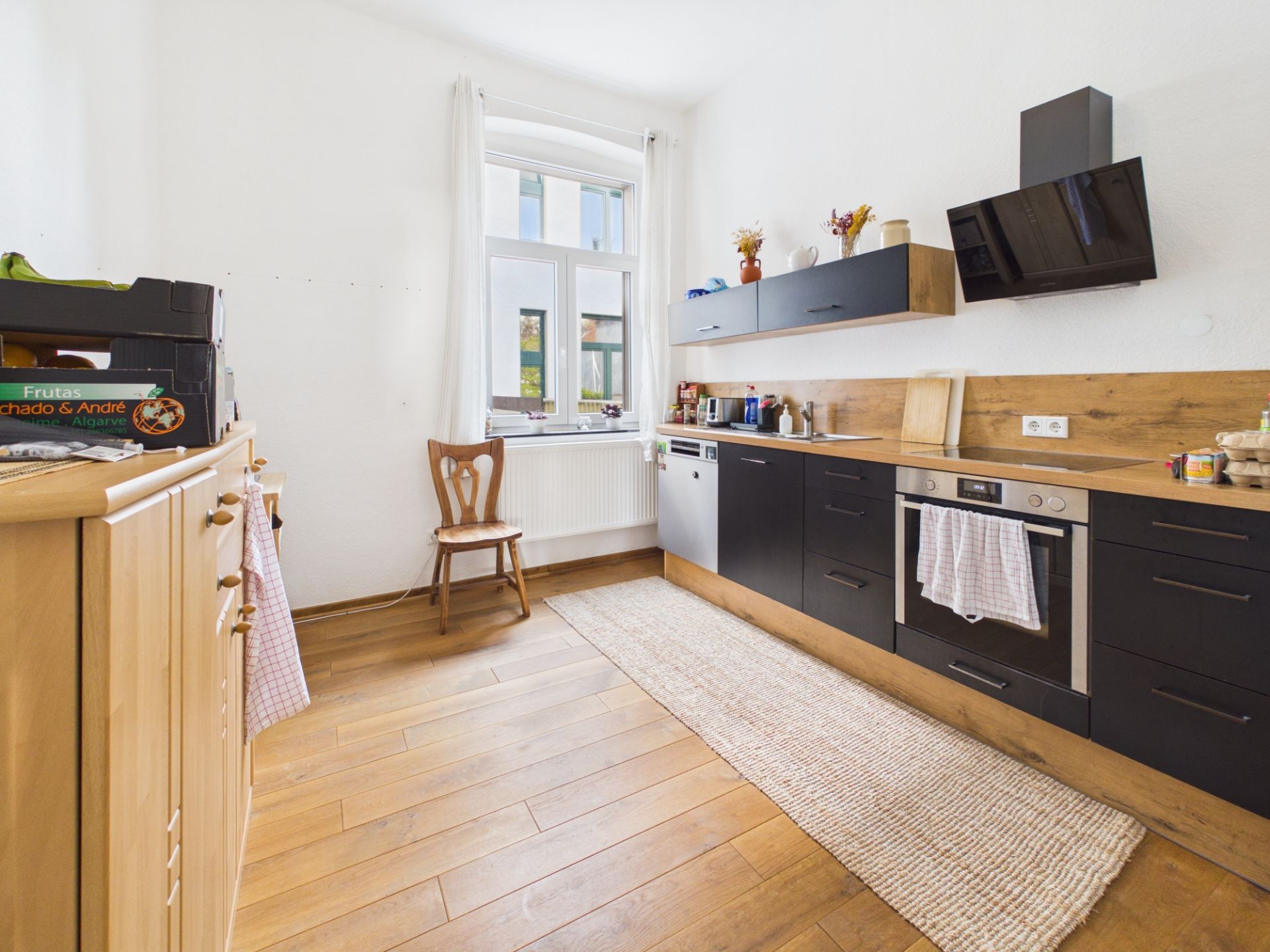 Helle Küche mit Holzfußboden, schwarzen Schränken, Ofen, Stuhl und Fenster.