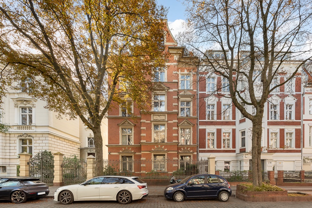 Mehrstöckiges Wohnhaus mit roter Fassade, Bäumen und geparkten Autos davor.