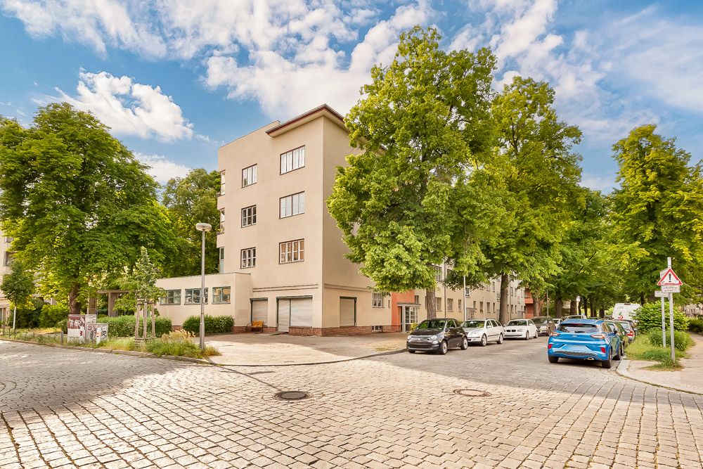 Mehrstöckiges Wohnhaus an einer ruhigen Straße mit parkenden Autos und vielen Bäumen.