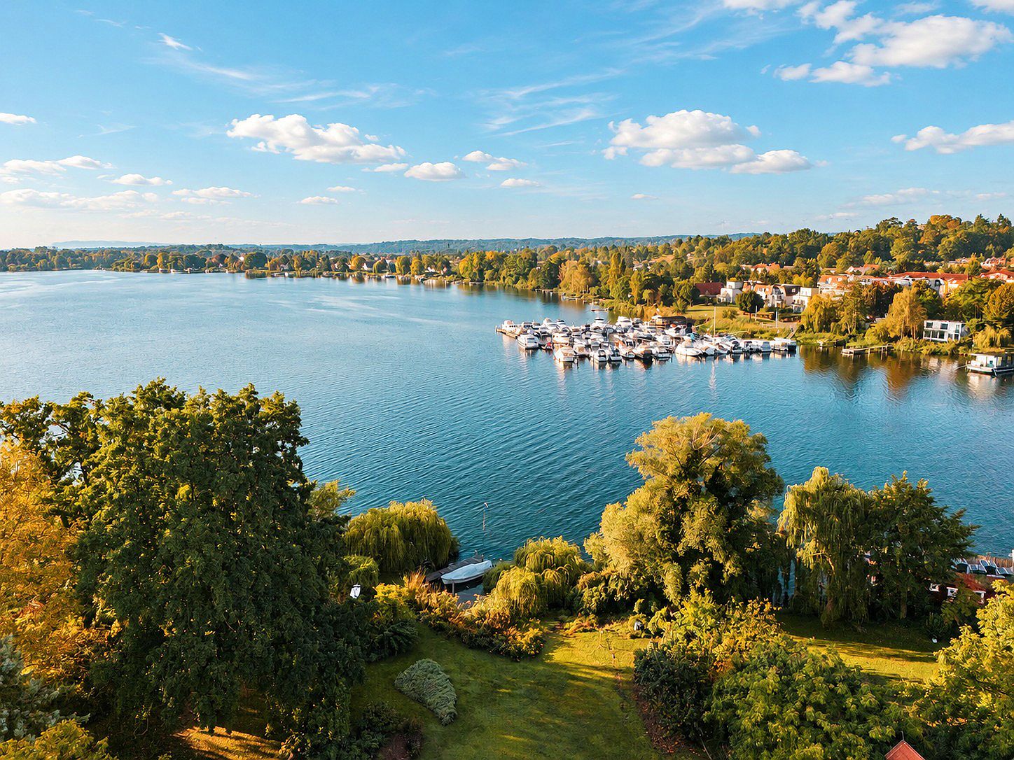 Blick auf einen See mit Booten, Bäumen und Häusern am Ufer bei sonnigem Wetter.