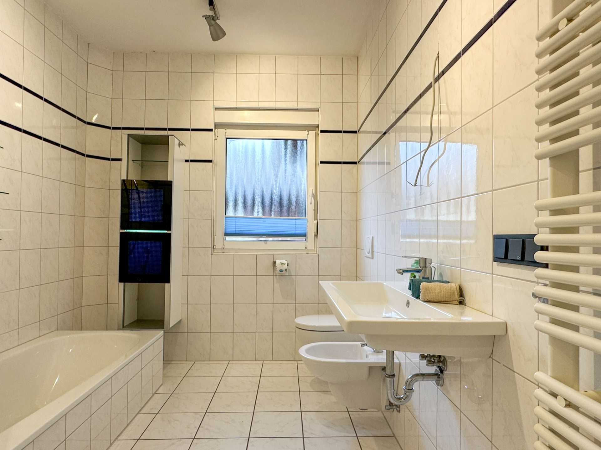 Helles Badezimmer mit Badewanne, Waschbecken, Bidet, Fenster und weiß gefliesten Wänden.