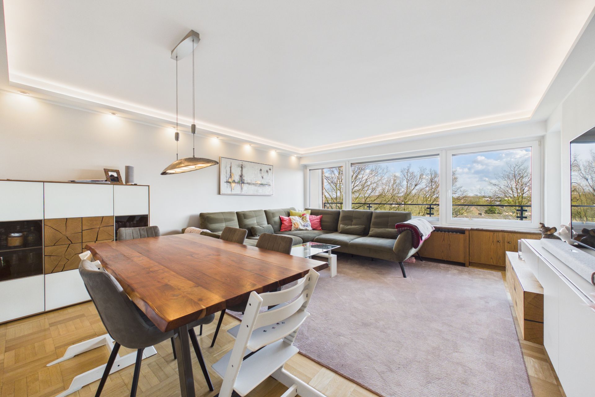 Helles Wohnzimmer mit großer Eckcouch, Esstisch und großen Fenstern mit Blick ins Grüne.