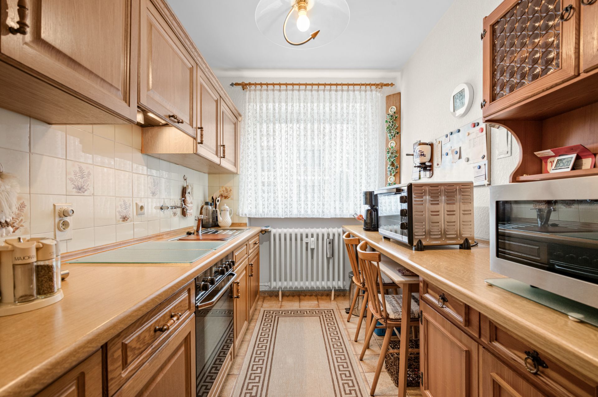 Kleine Küche mit Holzschränken, Elektroherd, Backofen und Fenster mit Gardine.
