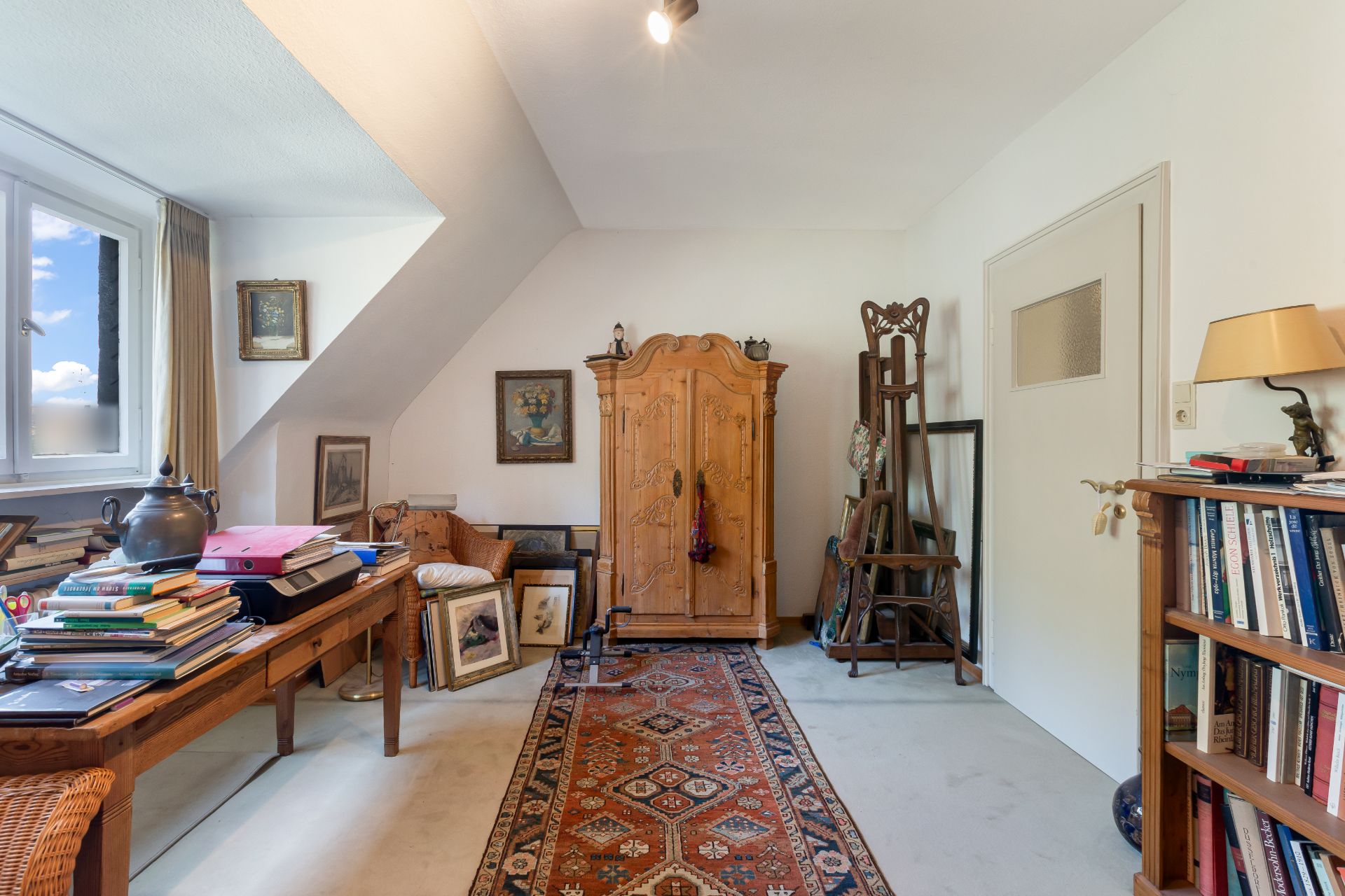 Wohnraum mit Holztisch, Teppich, Büchern, Schrank und Regal, Fenster links.