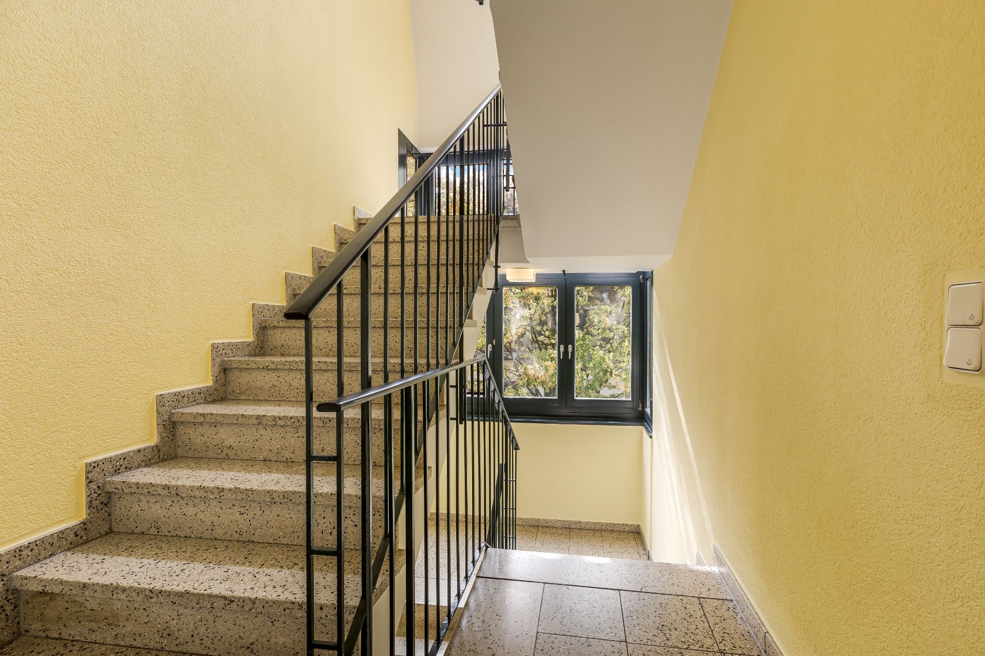 Helles Treppenhaus mit gelben Wänden, Fenstern und schwarzem Geländer.