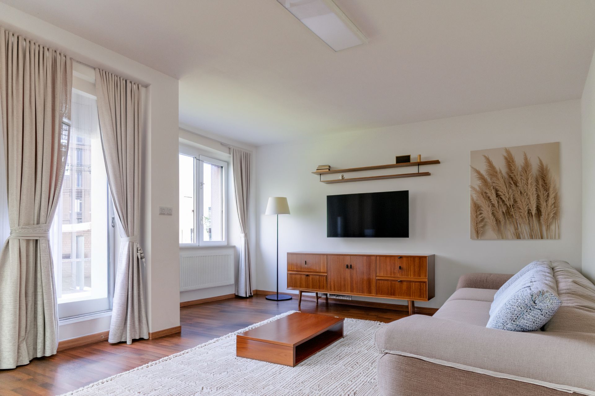 Helles Wohnzimmer mit Sofa, Couchtisch, Kommode, TV, Stehlampe und großen Fenstern mit Vorhängen.