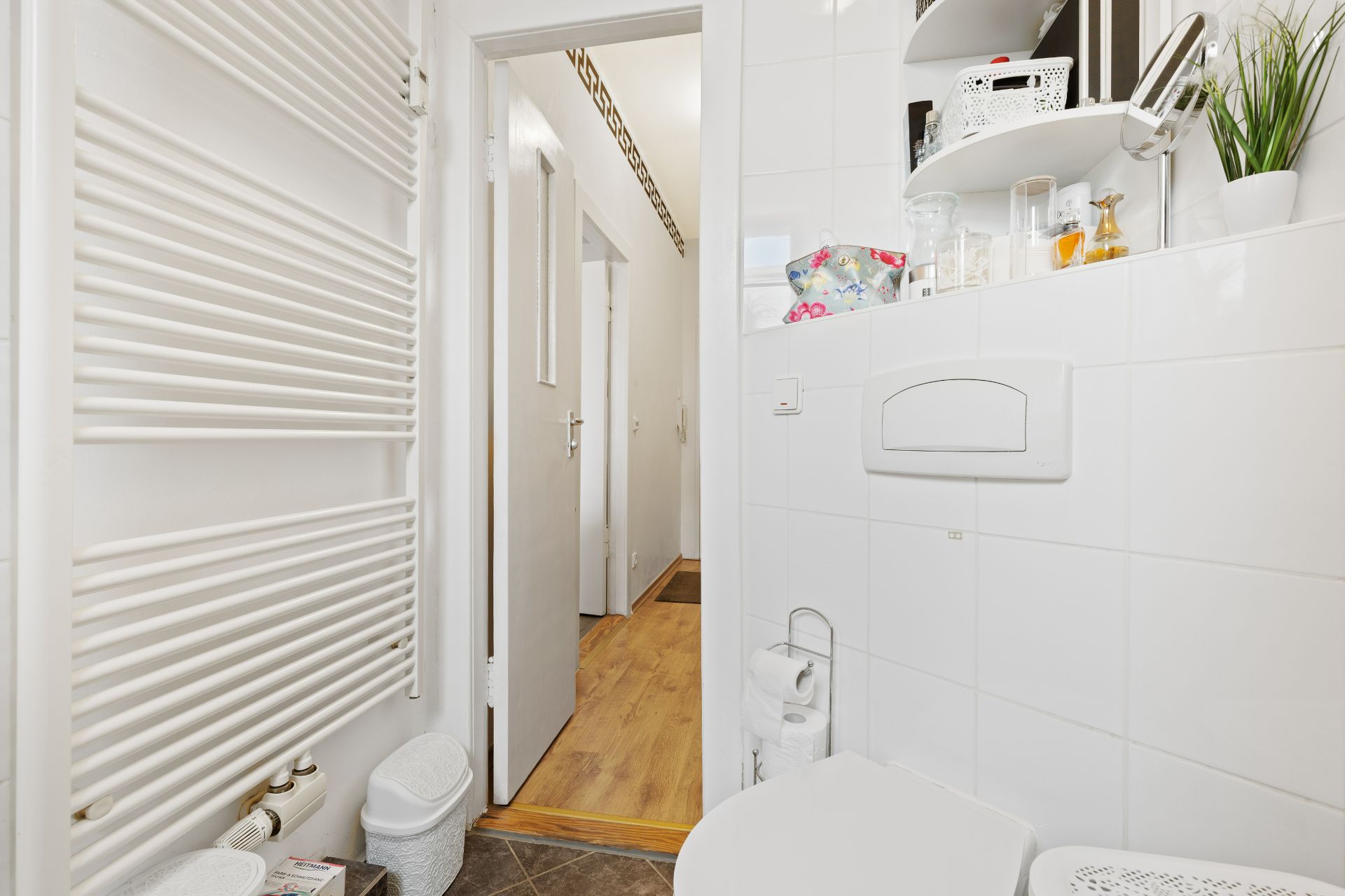 Helles Badezimmer mit Handtuchheizkörper, Toilette und Regal mit Utensilien.