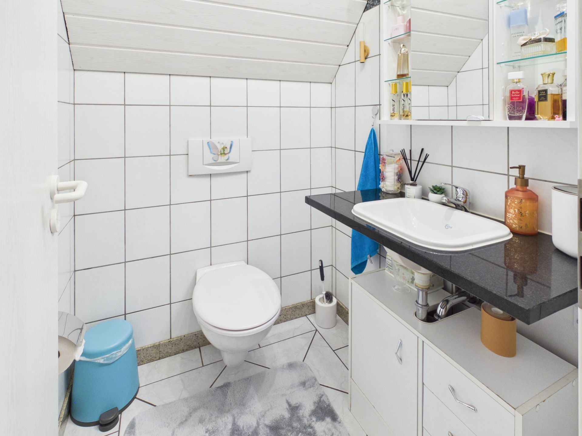 Kleines Badezimmer mit WC, Waschbecken, Regal und verschiedenen Pflegeprodukten.