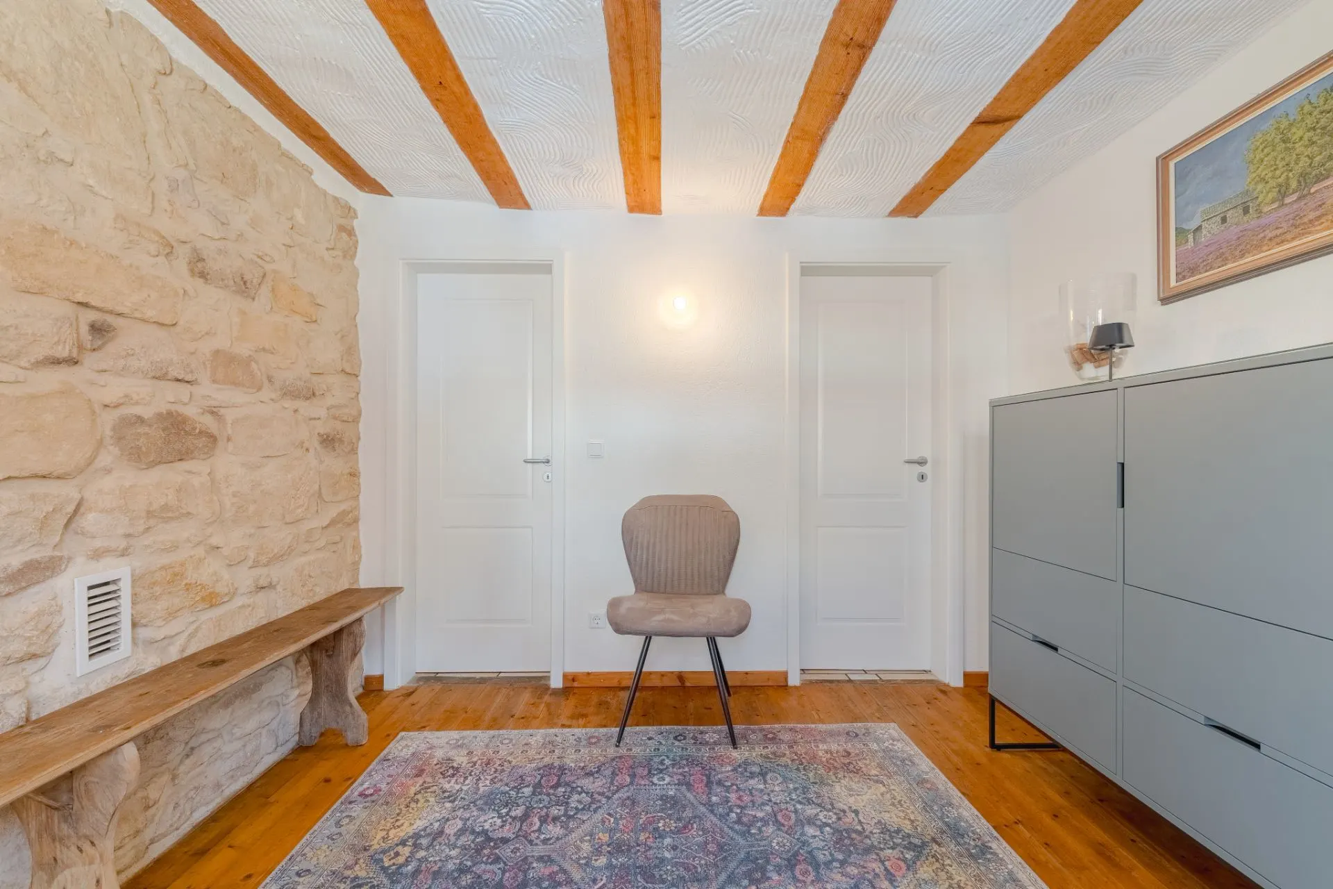 Zimmer mit Steinwand, zwei weißen Türen, Stuhl, Holzbank, Kommode und Teppich.