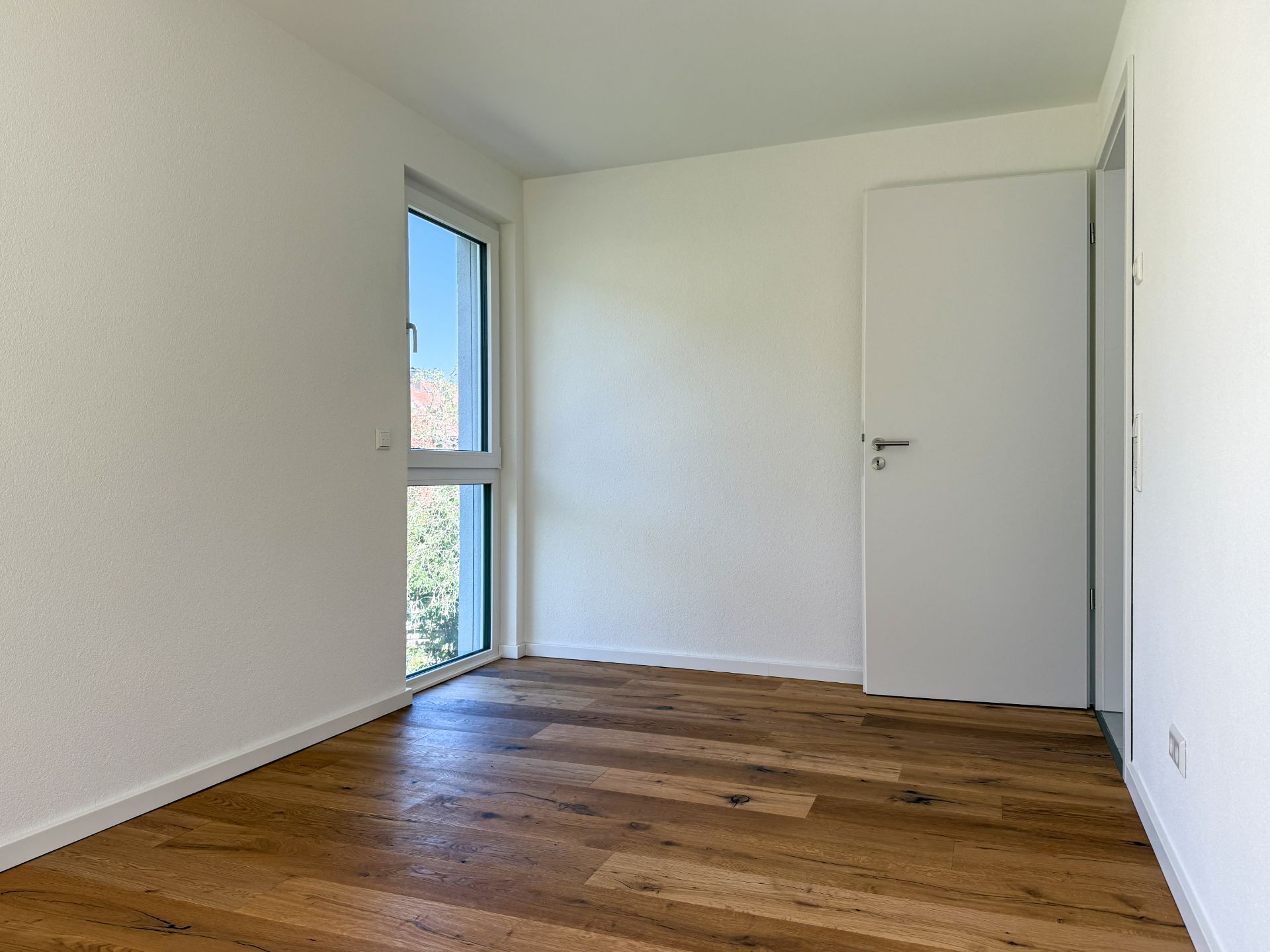 Leeres Zimmer mit Holzboden, weißer Wand, Tür und großem Fenster.