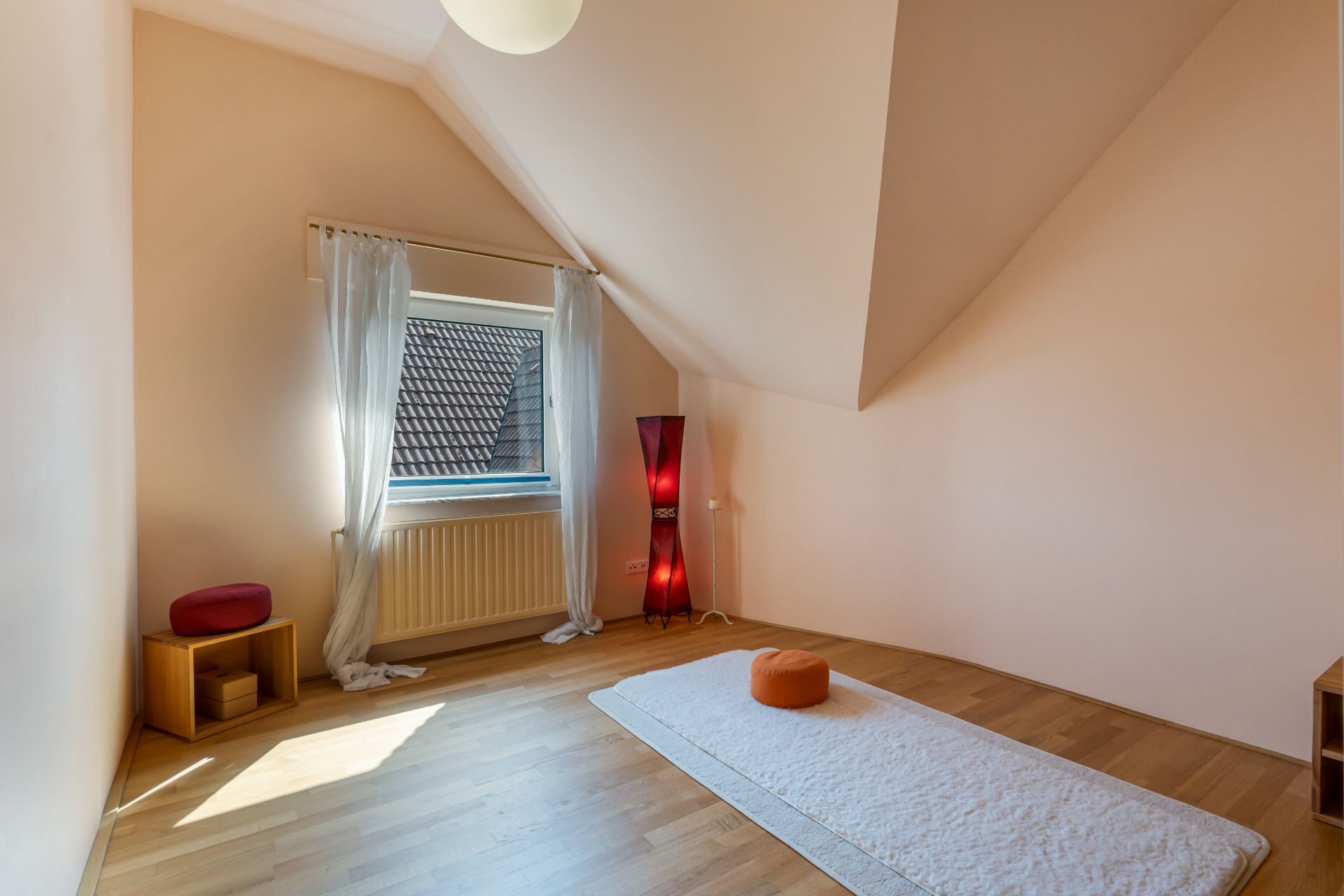 Helles Zimmer mit Holzfußboden, Fenster, weißem Teppich und orangenem Meditationskissen.