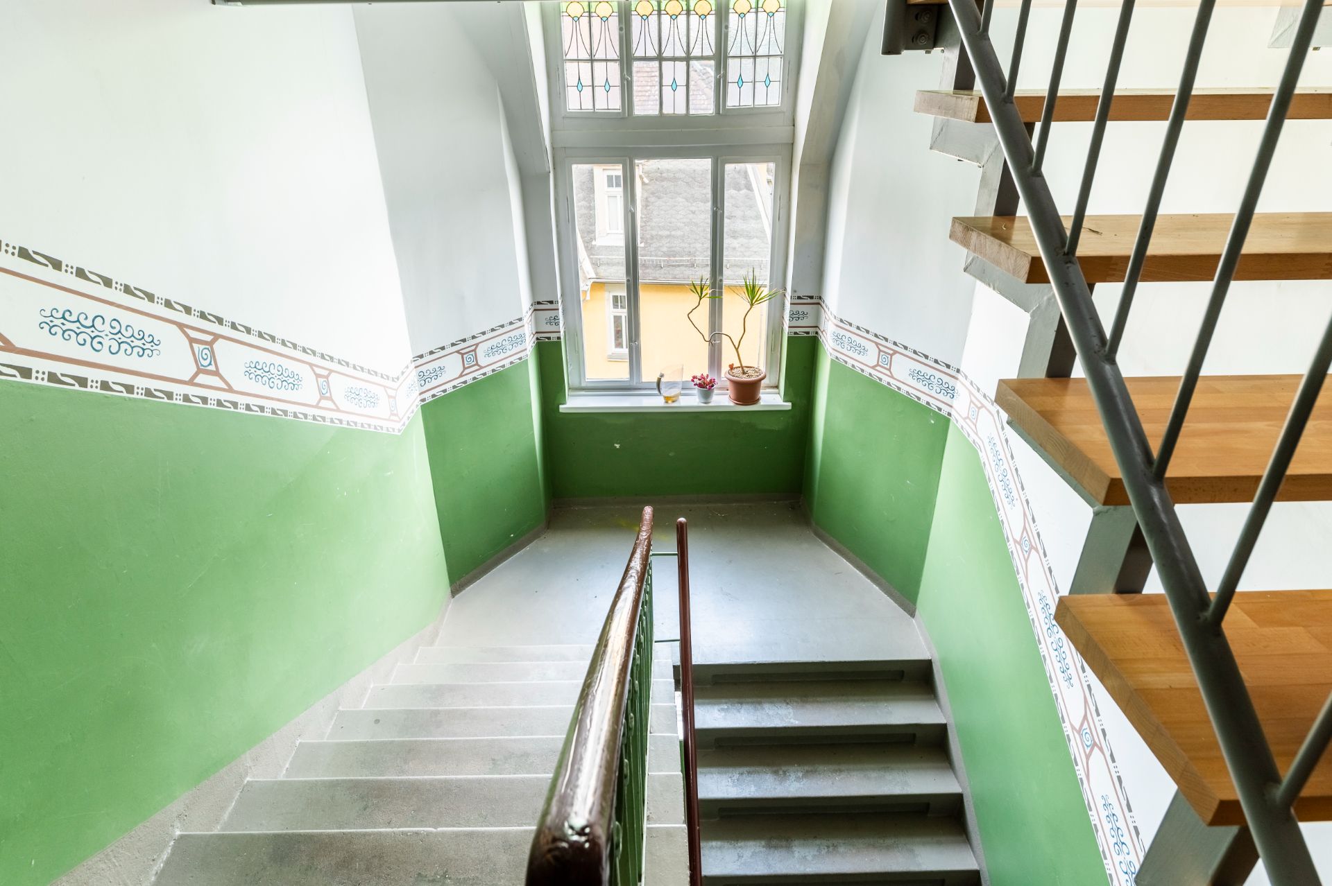 Treppenhaus mit grün-weißen Wänden, Holztreppe und Fenster mit zwei Blumentöpfen.