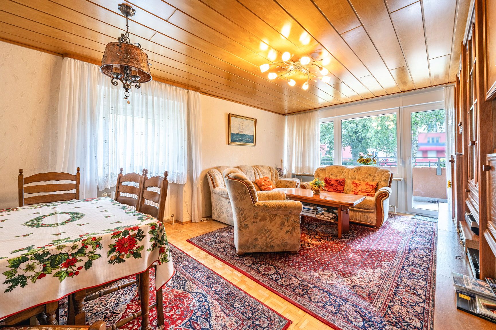 Wohnzimmer mit Sitzgruppe, Teppichen, Holzmöbeln und einem Esstisch mit Tischdecke.