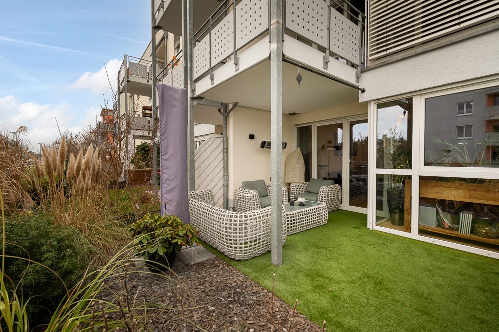 Überdachte Terrasse mit weißen Gartenmöbeln und Kunstrasen neben einem modernen Wohnhaus.