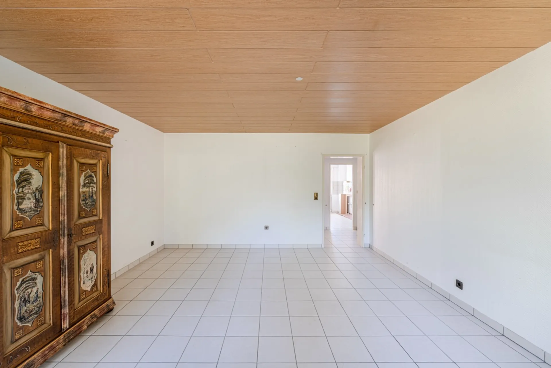 Leeres Zimmer mit Fliesenboden, Holzdecke und großem antiken Schrank links.
