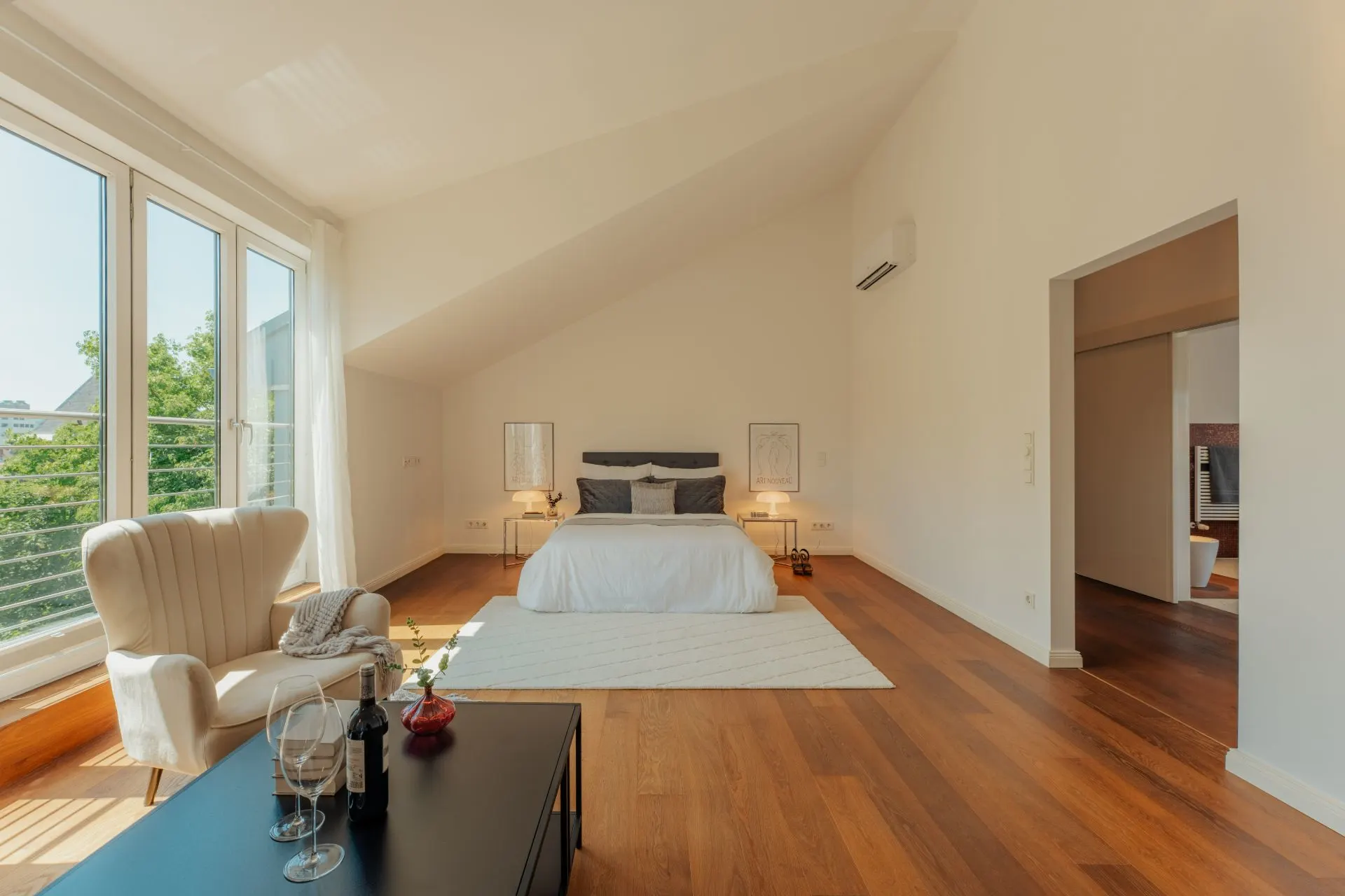 Helles Schlafzimmer mit großem Bett, Holzfußboden, Sessel und großen Fenstern.
