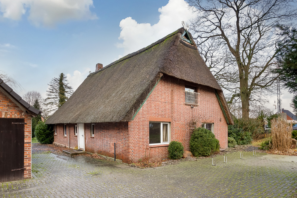 Backsteinhaus mit Reetdach, großen Fenstern und kleiner Terrasse, umgeben von Bäumen und Sträuchern.