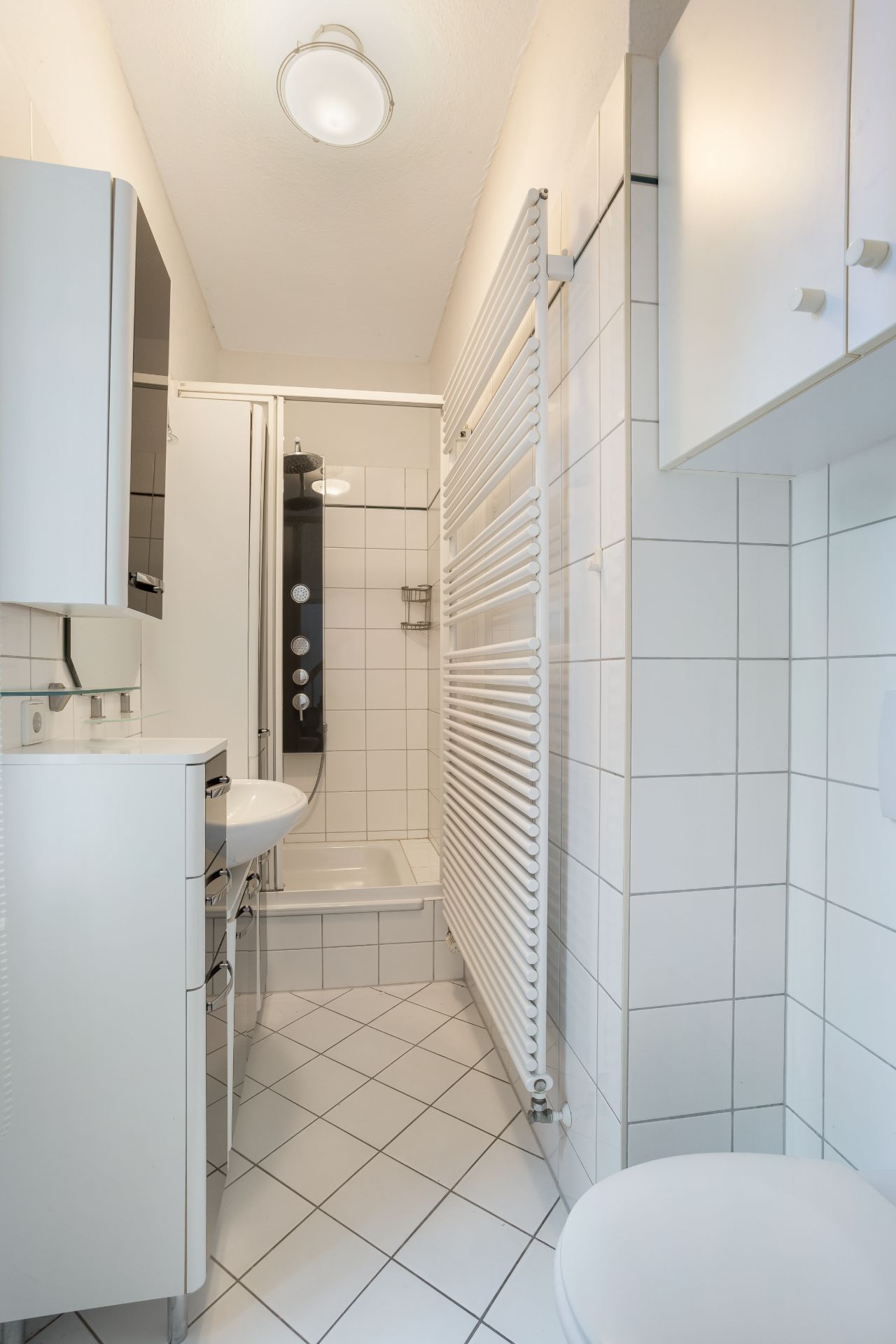 Kleines, weiß gefliestes Bad mit Dusche, Waschbecken, WC und Handtuchheizkörper.