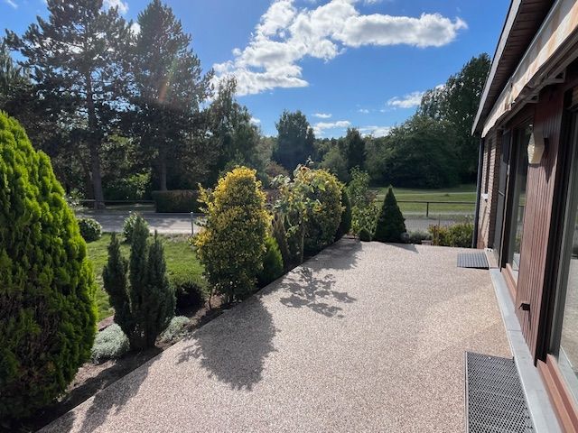 Sonnige Terrasse mit Kiesboden, Pflanzen am Rand und Blick auf einen Garten mit Bäumen.