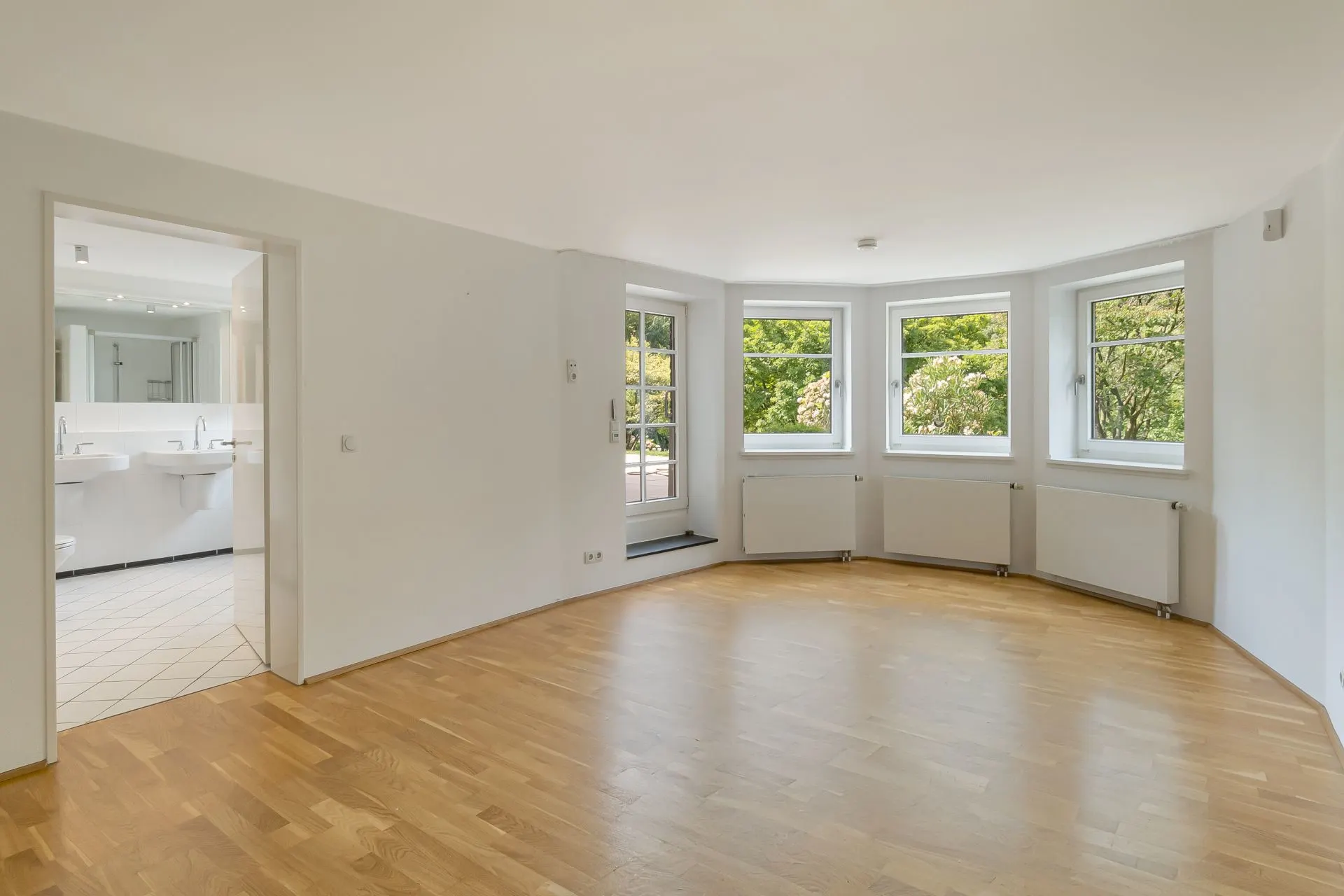 Helles Wohnzimmer mit Holzboden, mehreren Fenstern und Zugang zu einem angrenzenden Badezimmer.