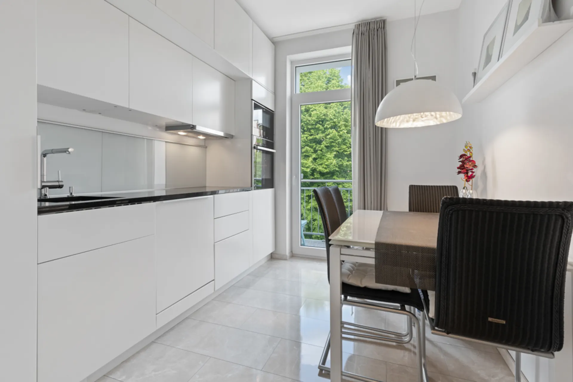 Moderne weiße Küche mit Esstisch, schwarzen Stühlen und großem Fenster mit Blick auf Bäume.
