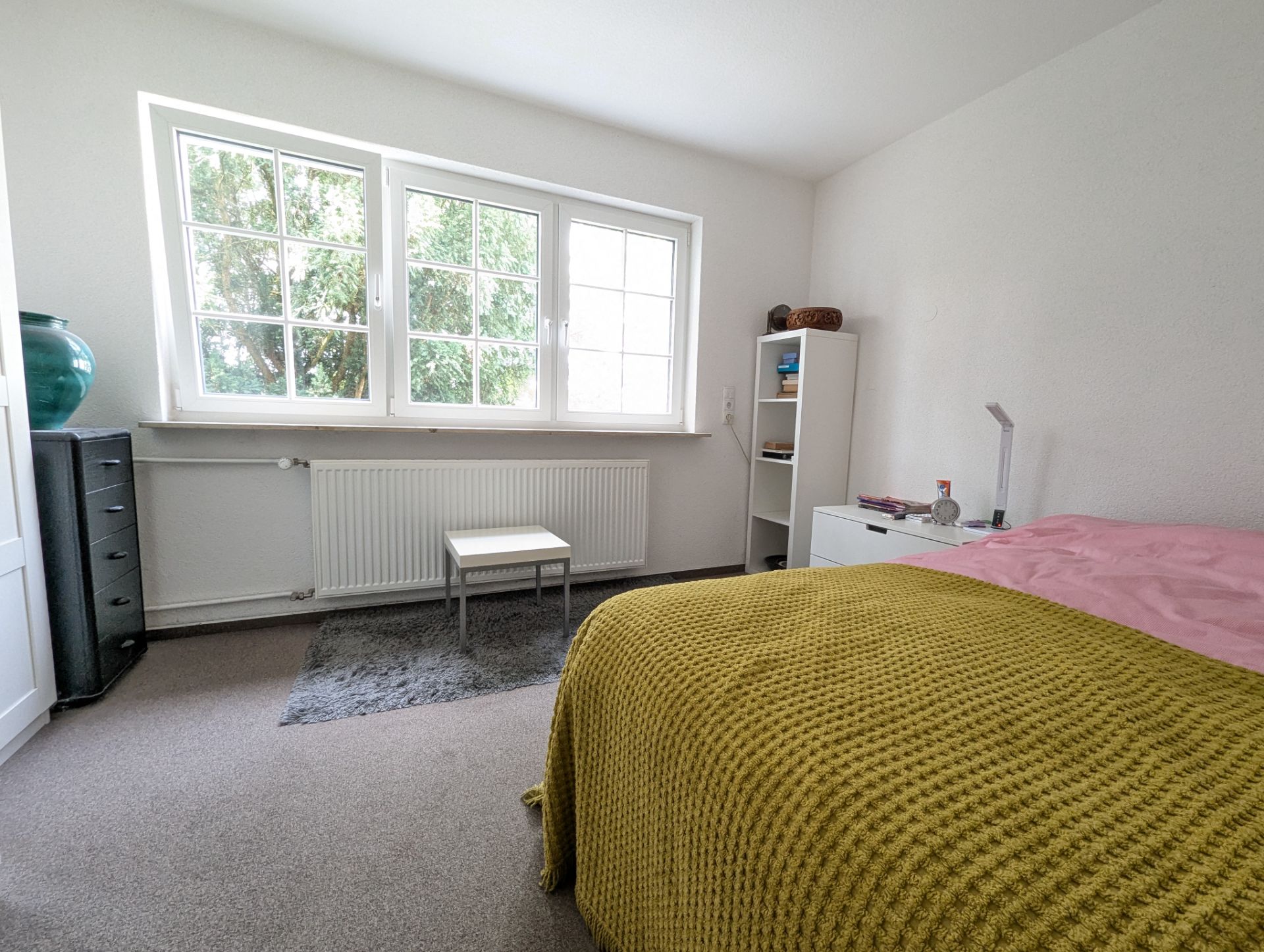 Helles Schlafzimmer mit großem Fenster, Bett, Regal und kleinem Tisch auf Teppich.