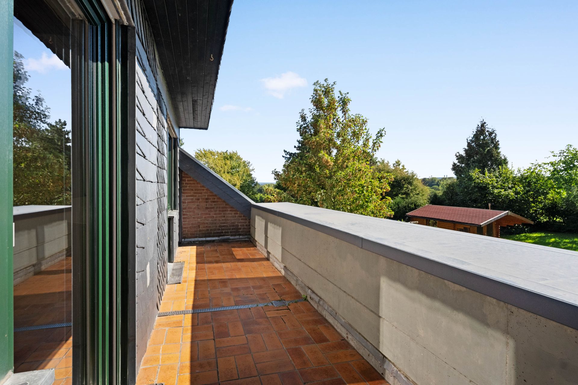 Balkon mit braunen Fliesen und Aussicht auf Bäume und Gartenhaus.