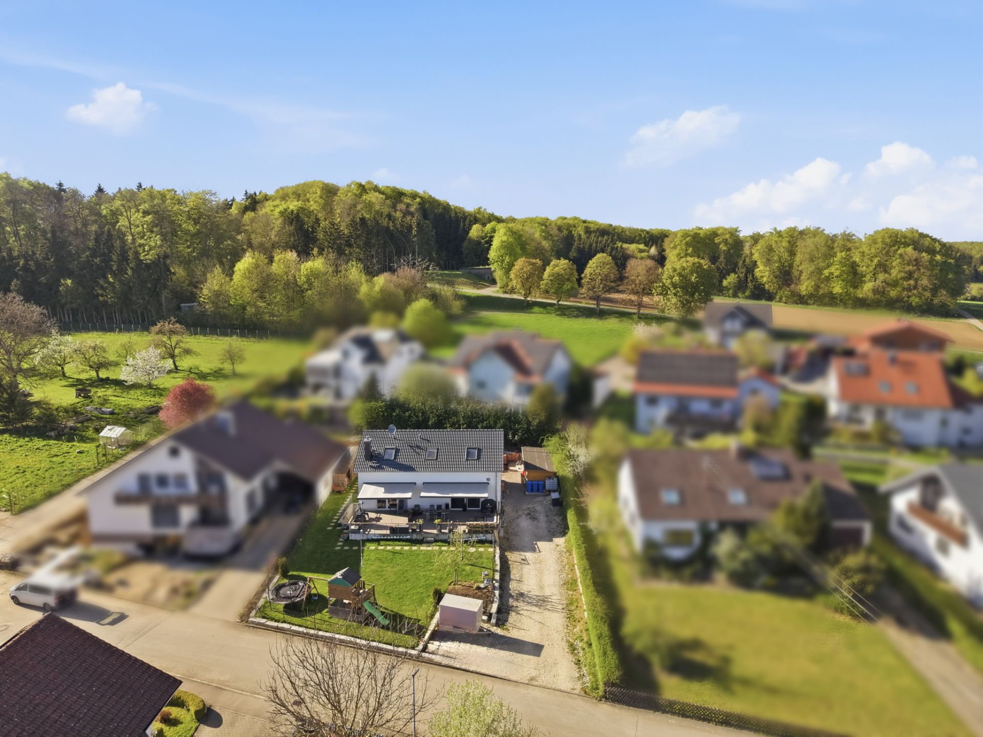 Luftaufnahme eines Wohnhauses mit Garten und umliegenden Häusern am Waldrand.