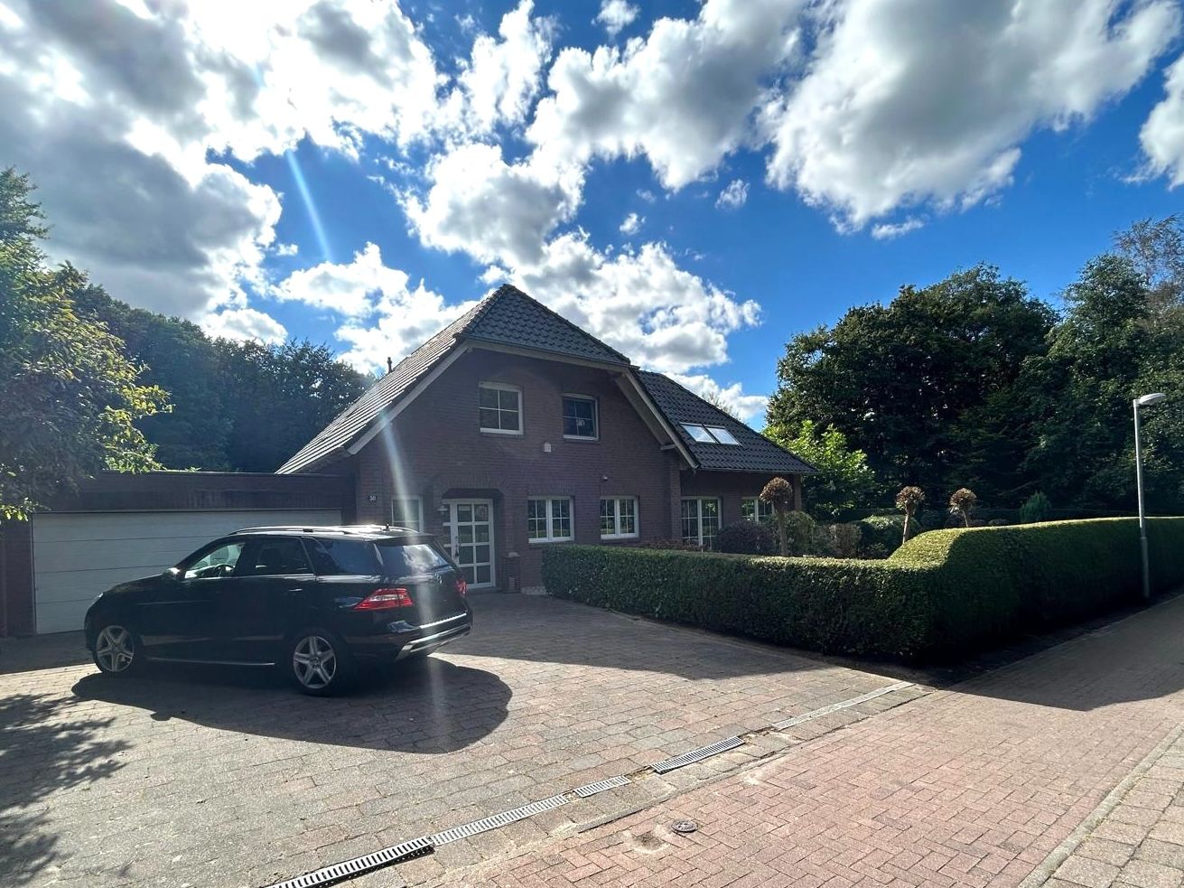 Backsteinhaus mit schwarzem Auto auf gepflasterter Einfahrt, umgeben von Hecken und Bäumen.
