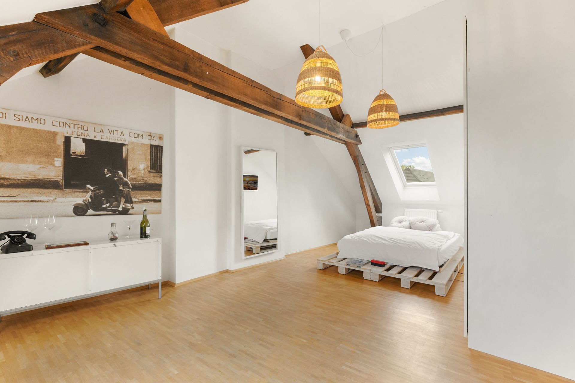 Helles Schlafzimmer mit Holzbalken, Bett auf Paletten, großem Spiegel und Dachfenster.