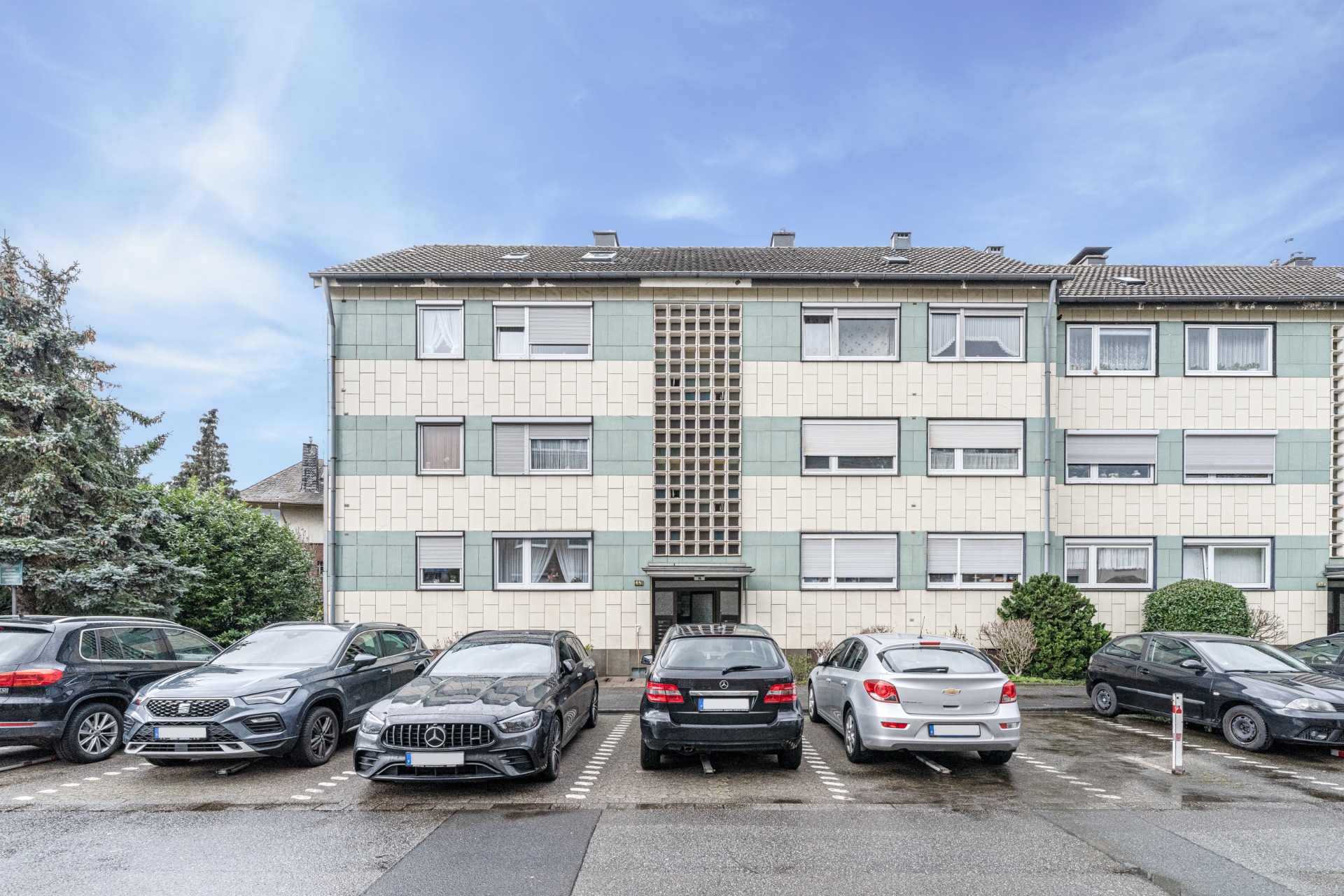 Dreistöckiges Wohnhaus mit geparkten Autos davor und bewölktem Himmel.
